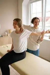 A therapist guides a woman in a stretching exercise on a massage table in a clinic setting.